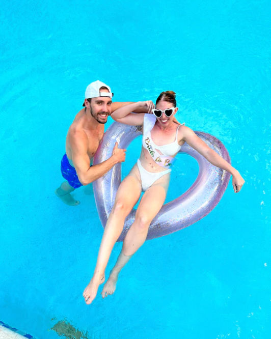 Smiling man in blue swim trunks and white cap gives a thumbs up while a woman in a white bikini and heart-shaped sunglasses lounges on a pink heart-shaped pool float in bright blue water