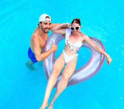 Smiling man in blue swim trunks and white cap gives a thumbs up while a woman in a white bikini and heart-shaped sunglasses lounges on a pink heart-shaped pool float in bright blue water