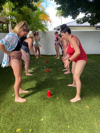 Women in swimsuits, barefoot and lined up facing each other, playing a cup game with red plastic cups on green turf in a sunny tropical backyard with palm trees and a white wall.