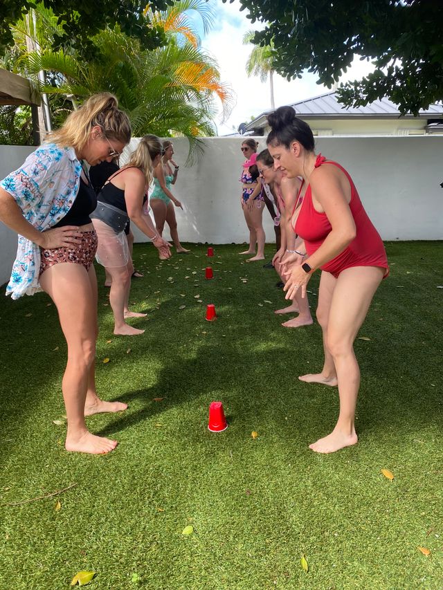 Group of women in swimsuits playing a red plastic cup relay game barefoot on green turf in a sunny tropical backyard with palm trees