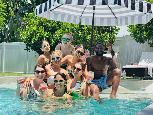 Eight friends smiling at a sunny backyard pool party under a black-and-white striped umbrella, wearing sunglasses and swimsuits and holding colorful drinks amid lush tropical greenery.
