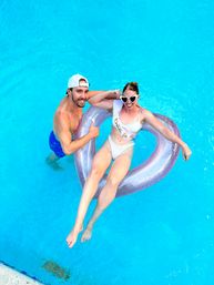 Overhead shot of a couple in a bright blue pool: woman in a white swimsuit and 'Bride to Be' sash lounging in a glittery heart-shaped float with heart sunglasses while a man in a white cap and blue trunks holds the float and gives a thumbs-up.