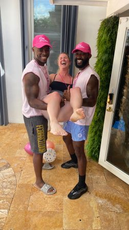 Two men wearing pink caps and matching shirts lift a laughing woman in a black swimsuit on a sunlit tiled patio with party balloons and greenery nearby.