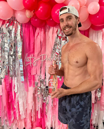Shirtless man wearing a white cap and camo shorts smiles while holding a sparkling rosé bottle in front of a pink balloon garland and fringe backdrop with a "Let's Party" sign.