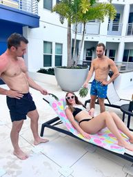 Three friends at a sunny tropical pool deck: a woman in a black bikini and heart-shaped sunglasses lounges on a colorful towel-covered chaise while two shirtless men in swim trunks playfully fan her with palm leaves near potted palms and a modern white building.