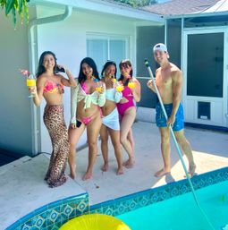 Smiling group of five friends at a sunny backyard pool: four women in colorful swimwear holding tropical cocktails with garnishes and one shirtless man with a pool skimmer by a tiled pool edge and yellow inflatable float.