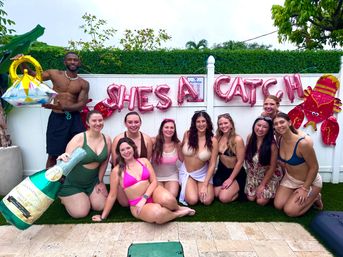 Poolside backyard bachelorette party: group of women in swimsuits and one man posing on a pool deck in front of pink "SHES A CATCH" balloons with oversized champagne and ring inflatables and tropical greenery