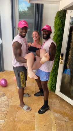 Sunny patio party: two men in pink caps and bead necklaces lift a laughing woman in a black swimsuit by a sliding glass door on stone tile with a hanging green plant.