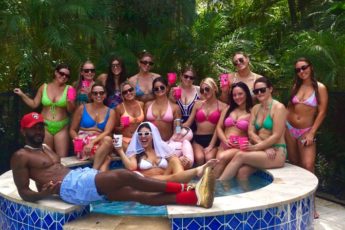 Tropical pool party group in a backyard hot tub: bride-to-be with veil and sunglasses surrounded by friends in colorful bikinis and one man lounging on the tub edge, everyone holding bright cups against a palm-leaf backdrop.