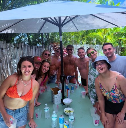 Smiling friends at a sunny tropical backyard pool party under a large umbrella, drinks on a table with palm trees and bamboo fence in the background