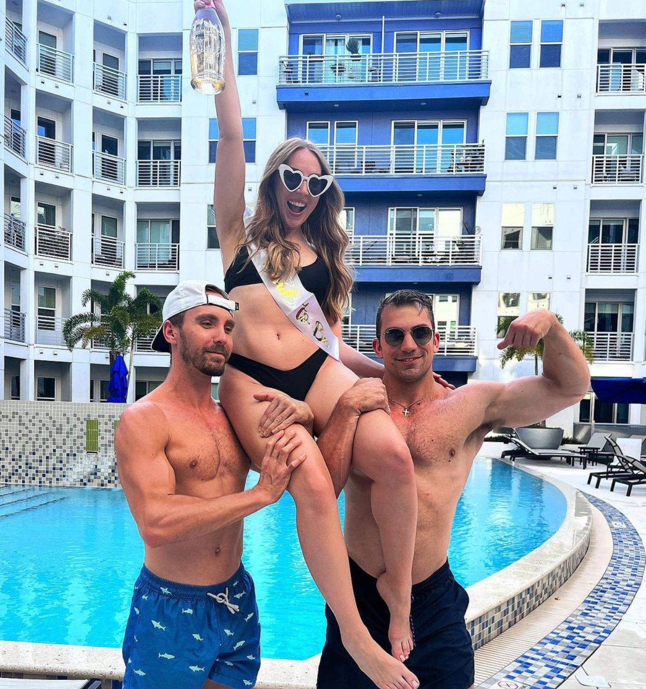 Woman in black bikini and heart-shaped sunglasses wearing a sash and raising a bottle while two shirtless men lift her at a resort-style apartment pool