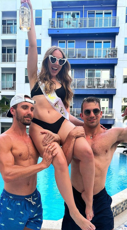 Woman in black bikini and heart-shaped sunglasses wearing a sash and raising a bottle while two shirtless men lift her at a resort-style apartment pool