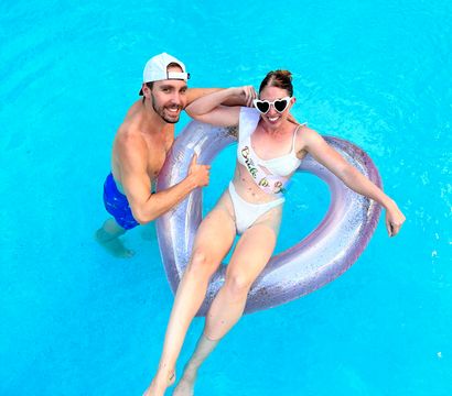 Aerial view of a smiling couple in a bright blue swimming pool: a woman in a white bikini and heart-shaped sunglasses lounges on a glittery heart-shaped inflatable wearing a "Bride to Be" sash while a man in blue swim trunks and a white cap holds the float, summer bachelorette pool celebration.