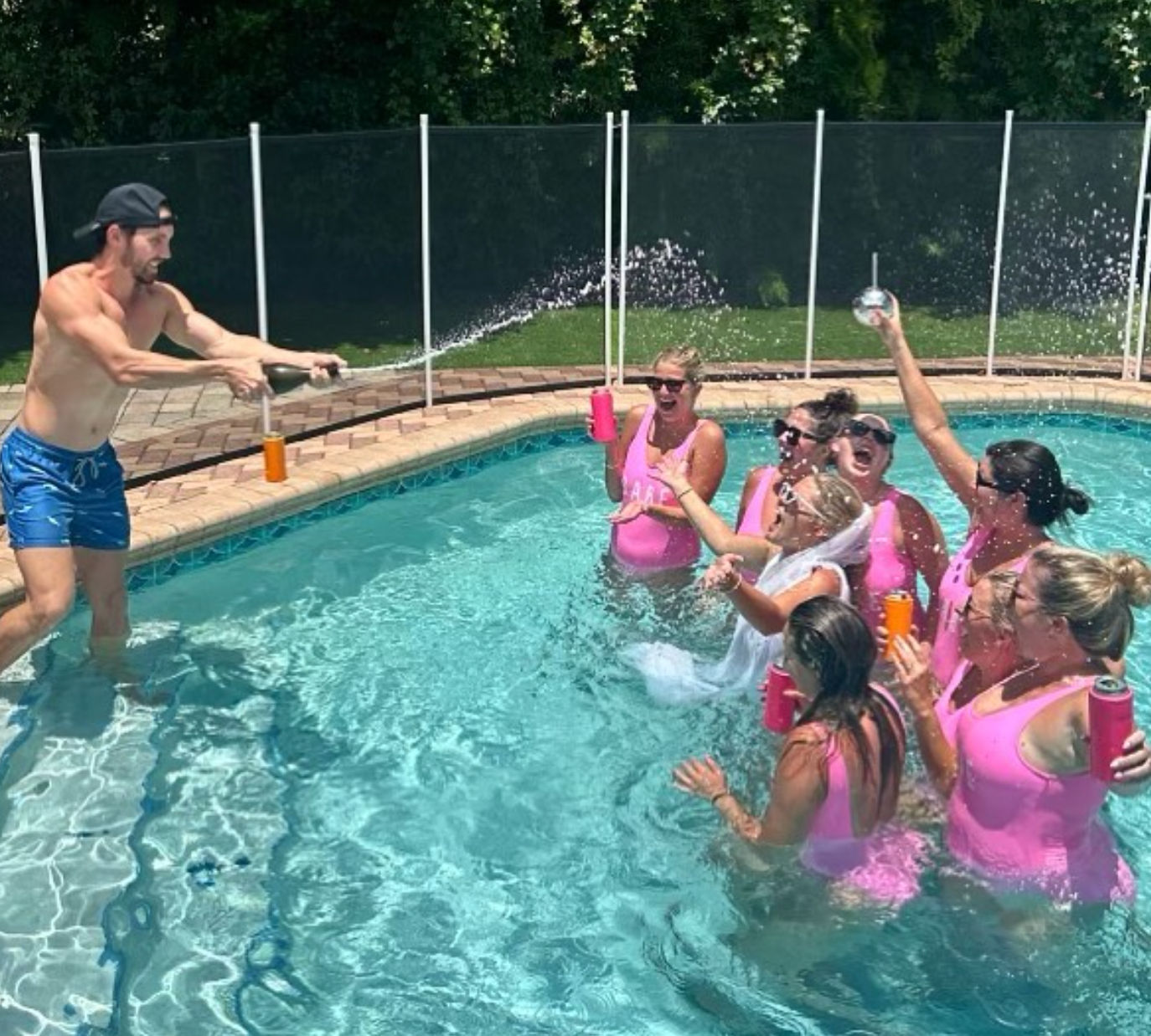 Man spraying champagne at a backyard pool as a group of women in matching pink swimsuits, including a veiled bride, cheer and splash with colorful tumblers.