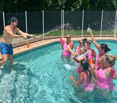 Man spraying champagne at a backyard pool as a group of women in matching pink swimsuits, including a veiled bride, cheer and splash with colorful tumblers.