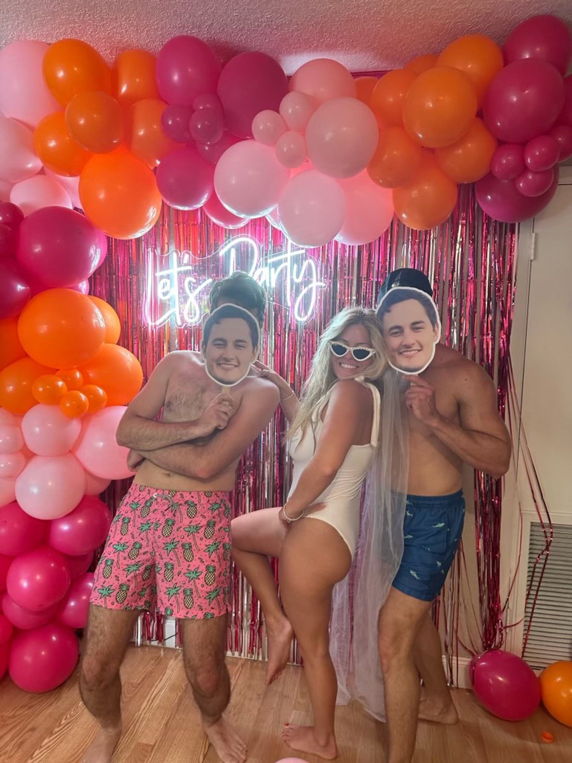 Three friends in swimwear pose under a pink-and-orange balloon arch and metallic fringe backdrop with a neon party sign; two hold face-cutout masks while a woman in a white swimsuit wears a veil and cat-eye sunglasses for a playful bachelorette-party vibe.