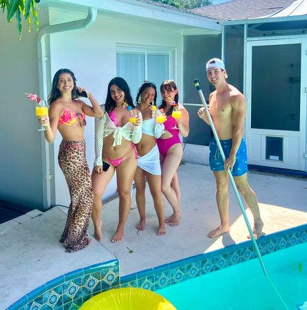 Five friends at a sunny backyard pool party – four women in colorful swimsuits holding tropical cocktails and a man in swim trunks with a pool skimmer beside a yellow float.