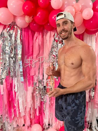 Smiling shirtless man in a backward white cap holding a rose-gold champagne bottle in front of a pink balloon garland and silver fringe party backdrop — indoor celebration.