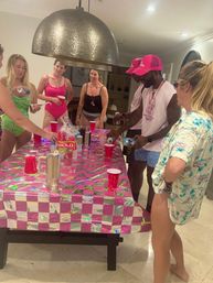 Indoor house party with friends in swimsuits gathered around a pink holographic checkerboard tablecloth, red Solo cups and drinks, some wearing pink caps and beads under a large pendant light.