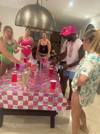 Indoor beach-party scene with people in swimsuits and pink hats gathered around a pink checkered table covered with red Solo cups and a large pendant light overhead.