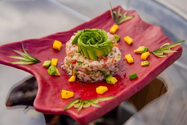 Seafood ceviche tartare crowned with an avocado rosette, black sesame, mango and cucumber garnishes on a vibrant red plate.