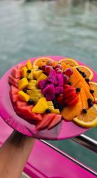 Vibrant pink plate of assorted tropical fruit — watermelon, mango, pineapple, dragon fruit, strawberries, grapes, cantaloupe, orange slices and blueberries — held over turquoise water on a boat.