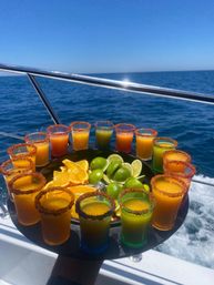 Tray of colorful chili-rimmed cocktail shots arranged around lime and orange wedges, held on a boat with sparkling blue ocean and clear sky in the background