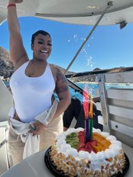 Smiling woman in a white swimsuit holding a towel, raising her arm aboard a boat next to a fruit-topped birthday cake with colorful sparkler candles, blue ocean and rocky coastline under a sunny sky.