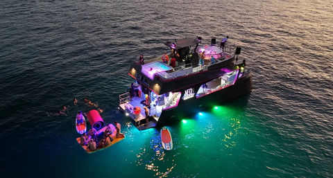 Neon-lit double-deck party boat anchored at sunset over turquoise sea, guests swimming around a floating platform and paddleboard.