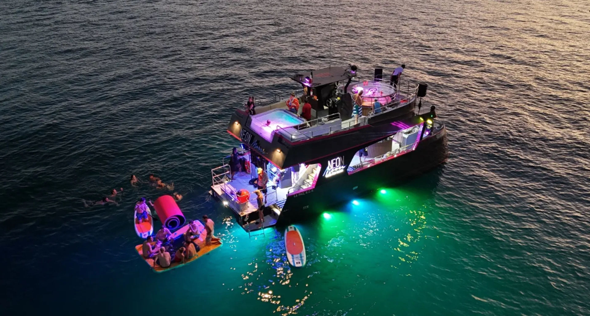 Neon-lit double-deck party boat anchored at sunset over turquoise sea, guests swimming around a floating platform and paddleboard.