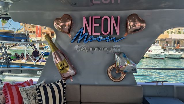 Party yacht interior at Cabo marina with a neon 'NEON Moon' sign reading 'Cabo made me do it', rose-gold heart and champagne balloons that say "let's party", a ring balloon, striped pillows, and boats visible through the window.