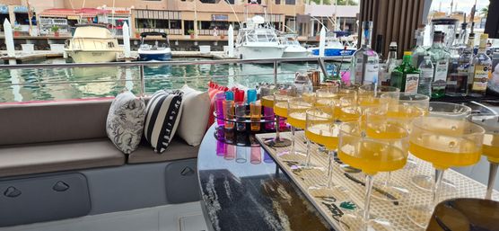 Yacht bar at a marina with cushioned bench and striped pillows, colorful shot glasses and rows of yellow cocktails on a glossy counter, boats and harbor in the background.