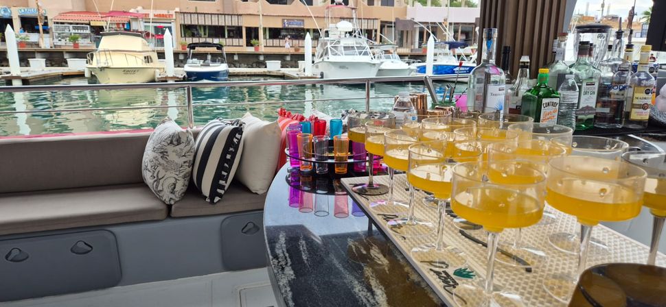 Yacht bar at a marina with cushioned bench and striped pillows, colorful shot glasses and rows of yellow cocktails on a glossy counter, boats and harbor in the background.