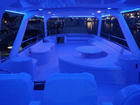 Luxury yacht deck at night with blue LED ambient lighting, curved white lounge seating and round table overlooking a lit marina and water reflections.