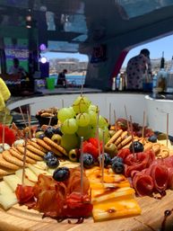 Close-up charcuterie board on a wooden platter with green grapes, blueberries, olives, crackers, sliced cheeses and rolled cured meats drizzled with balsamic, served aboard a yacht with marina and waterfront buildings in the background.