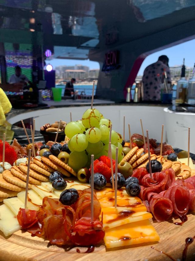 Close-up charcuterie board on a wooden platter with green grapes, blueberries, olives, crackers, sliced cheeses and rolled cured meats drizzled with balsamic, served aboard a yacht with marina and waterfront buildings in the background.