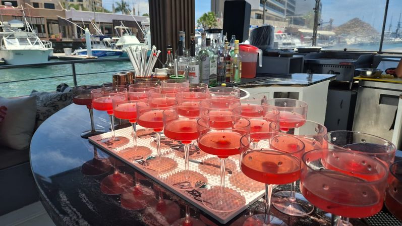 Sunlit row of bright red cocktails in coupe glasses on a boat bar, overlooking a marina with docked yachts and waterfront buildings.
