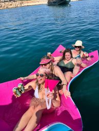 Four women in swimsuits laughing and holding drinks while lounging on a bright pink floating mat in clear blue ocean near a rocky shoreline and anchored boat.