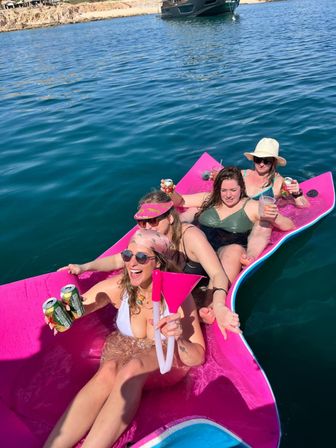 Four women in swimsuits laughing and holding drinks while lounging on a bright pink floating mat in clear blue ocean near a rocky shoreline and anchored boat.
