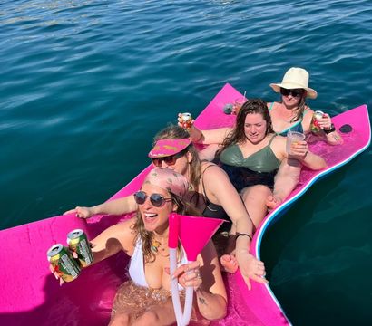 Four women in swimsuits laughing and holding drinks while lounging on a bright pink floating mat in clear blue ocean near a rocky shoreline and anchored boat.