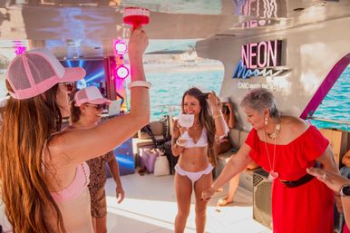 Bachelorette party on a sunny yacht — women in pink caps and swimsuits cheer with bright cocktails while the bride in a white bikini holds a 'Bride' goblet, turquoise ocean and neon party lights in the background.