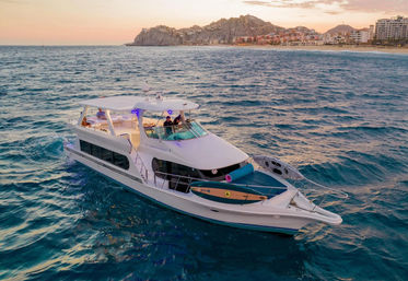 Luxury white yacht cruising turquoise ocean at sunset with a paddleboard on the bow and a beachfront resort and rocky headland on the distant coastline