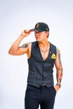 Studio portrait of a tattooed man tipping a black baseball cap while wearing a fitted black vest with a yellow pocket square and dark pants against a clean white background
