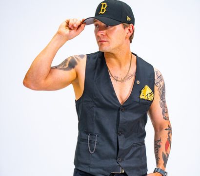 Studio portrait of a tattooed man tipping a black baseball cap while wearing a fitted black vest with a yellow pocket square and dark pants against a clean white background