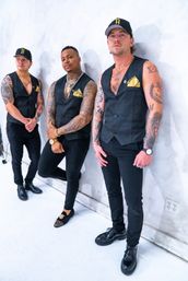 Three tattooed men in coordinated black vests, gold pocket squares and caps posing against a white studio wall — edgy urban menswear portrait.