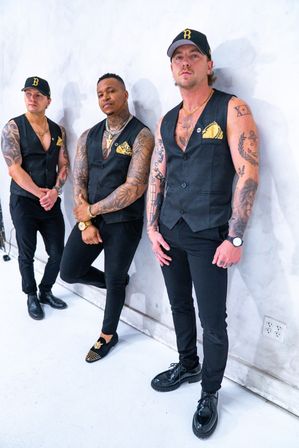 Three tattooed men in coordinated black vests, gold pocket squares and caps posing against a white studio wall — edgy urban menswear portrait.