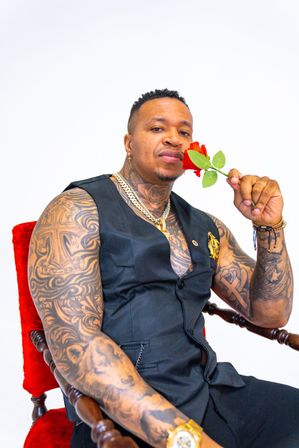 Bold tattooed man in a black vest and gold chains seated on a red velvet chair, holding a red rose to his face in a bright studio portrait
