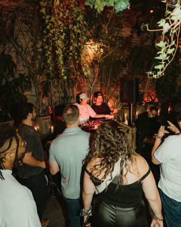 Crowd enjoying a lively evening DJ set at a plant-filled urban garden bar with hanging vines, warm candlelight, and tropical decor.