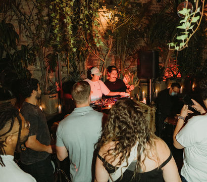 Crowd enjoying a lively evening DJ set at a plant-filled urban garden bar with hanging vines, warm candlelight, and tropical decor.