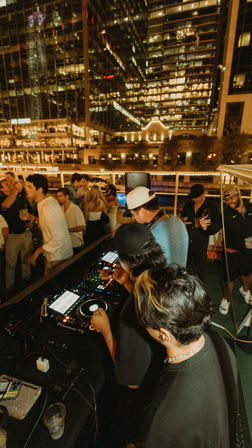 Nighttime boat party with DJs mixing at a deck DJ booth and a lively crowd, glowing downtown skyscrapers and reflections lighting the waterfront